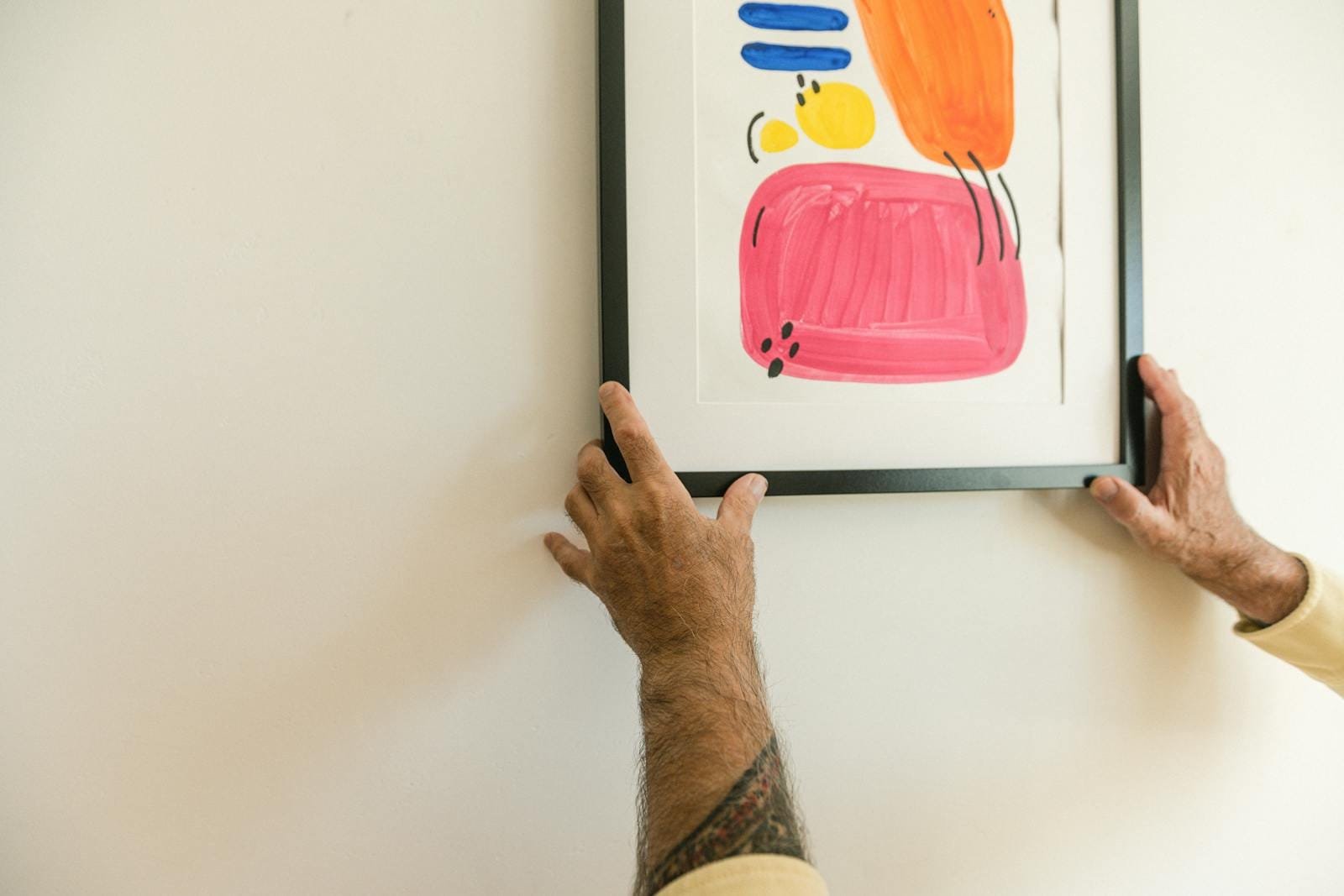 Two tattooed hands hang a colorful abstract framed artwork on a white wall indoors.